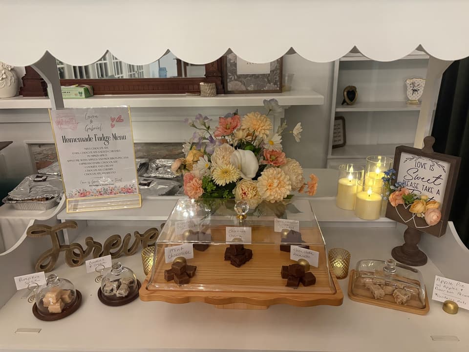 Homemade fudge pieces arranged beneath a glass cover among flowers, candles, and a wedding cart menu
